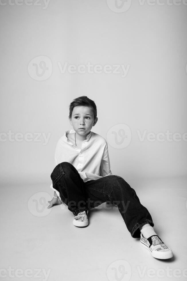 Black and white studio portrait of a young boy standing alone, arms relaxed, gazing slightly to the side. Simple backdrop and soft lighting create a calm, timeless mood. photo