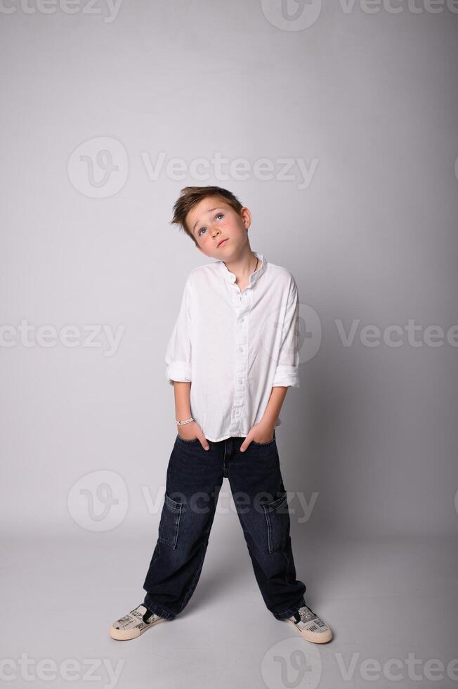Monochrome image of a young boy standing barefoot on a plain backdrop, arms crossed, with a confident and slightly defiant look. Stark lighting adds depth and contrast. photo