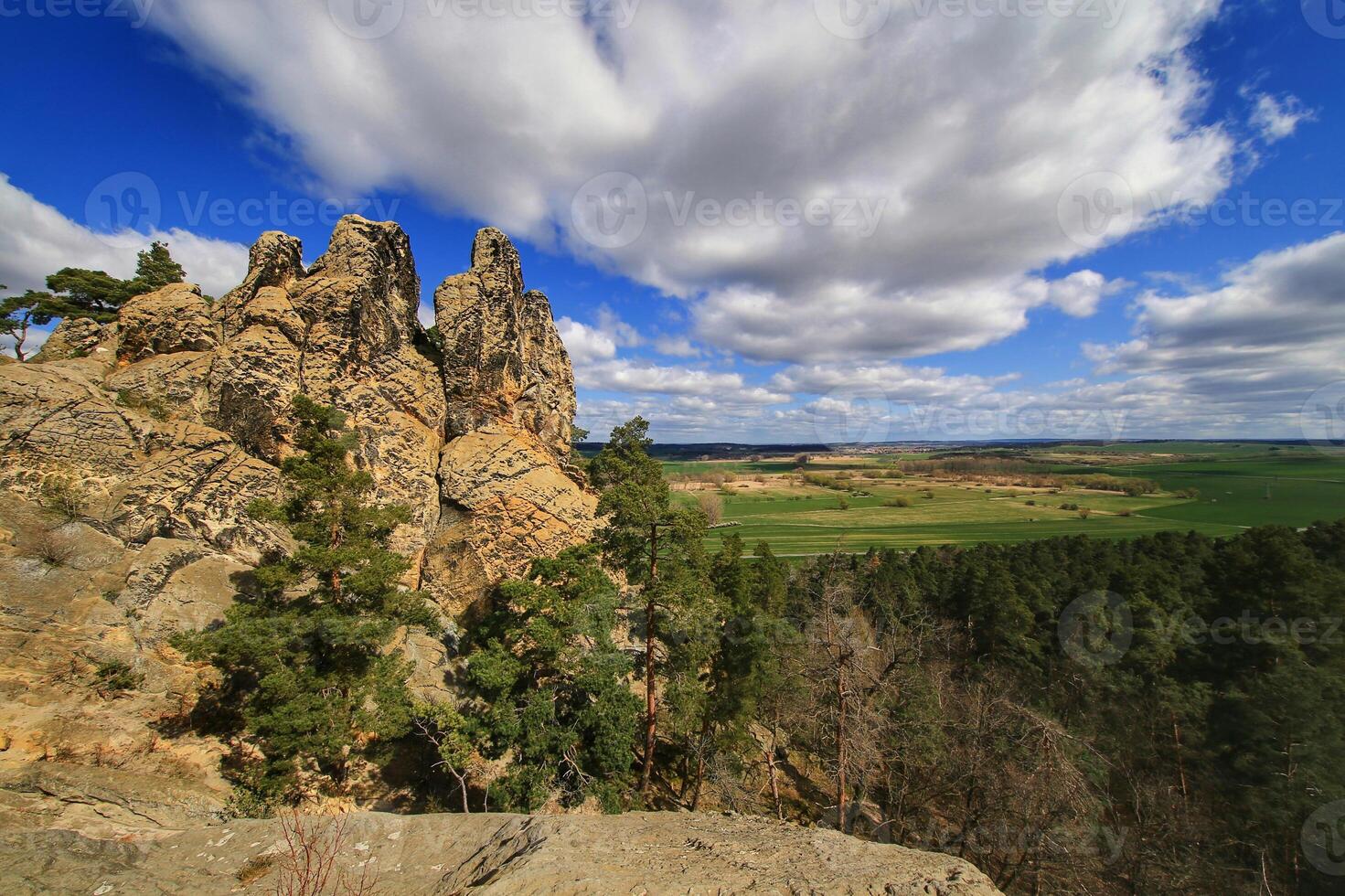 Sandstone spires stand above forested slopes and open farmland beneath rolling clouds photo
