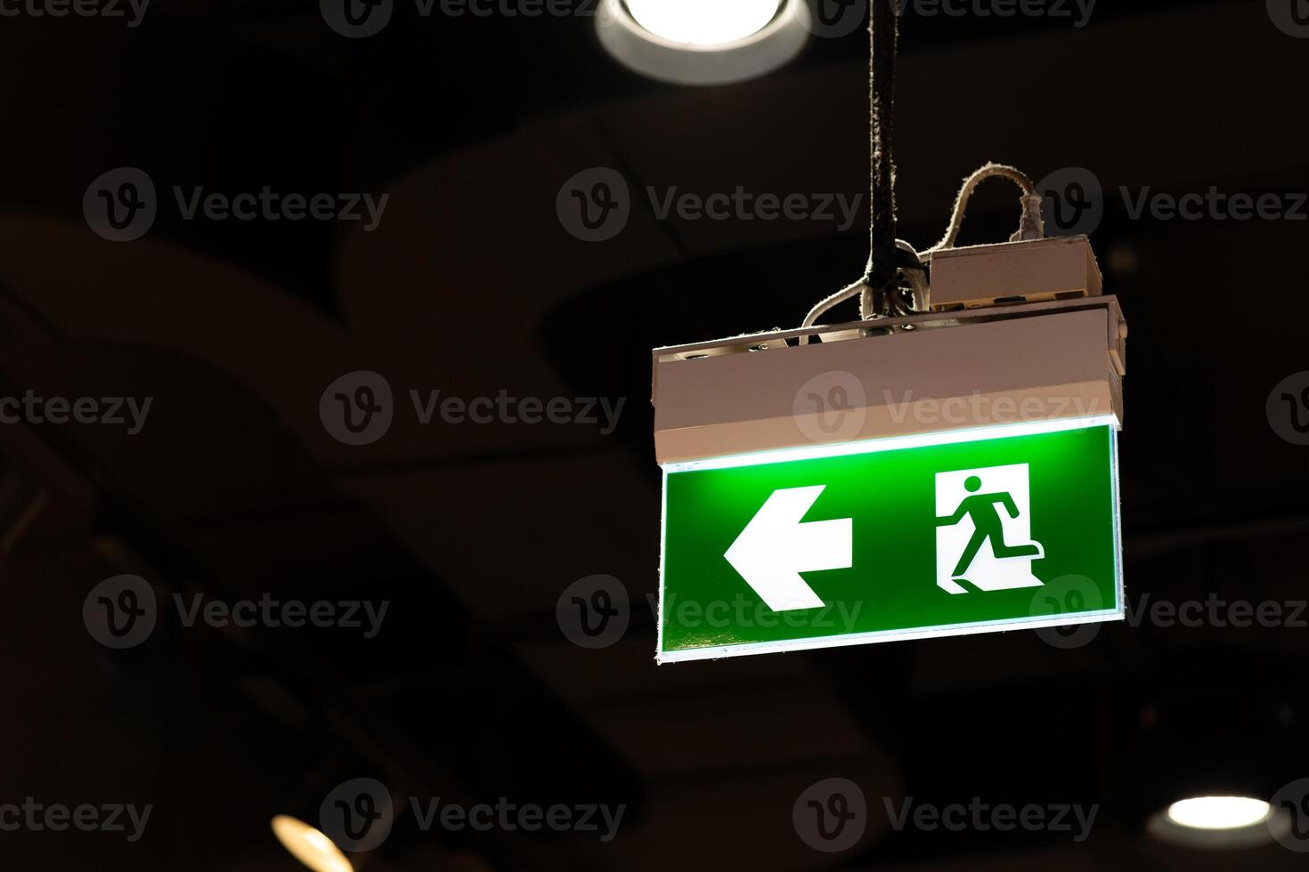 Illuminated Green Emergency Exit Sign with Arrow Pointing Left. green emergency exit sign hangs from the ceiling in a dimly lit indoor space. concepts of safety, evacuation, emergency preparedness. photo