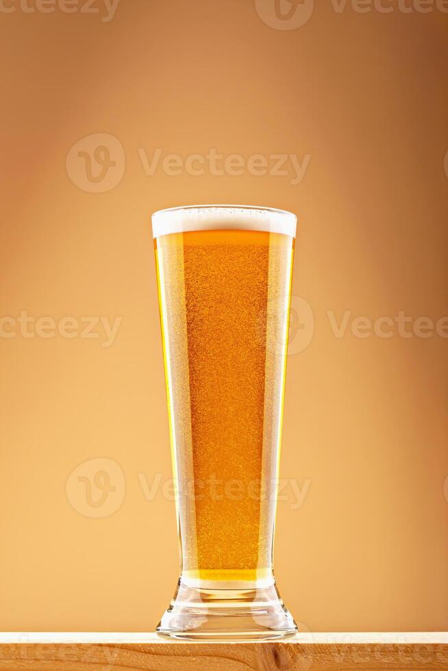 Pint of bright golden beer with light foam on wooden plank in warm ambient light. Concept of beer refreshment and lager clarity. Tall glass of golden beer with foam on top on wooden surface photo