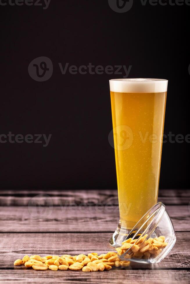 Tall glass of cold beer with frothy head beside spilled peanuts on wooden surface with black background. Concept of bar snack, beer pairing and casual pub moment. Glass of beer with peanuts on wood photo
