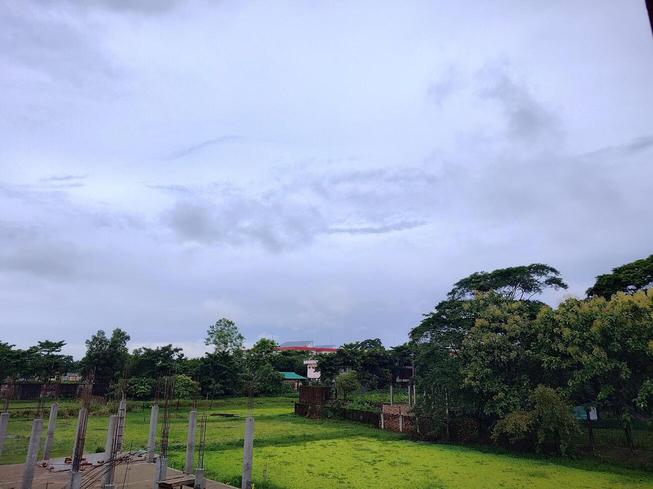 Overcast outdoor scene. Green grass, concrete pillars, rebar construction. Lush trees, buildings, and a red roofed house define this suburban landscape. photo