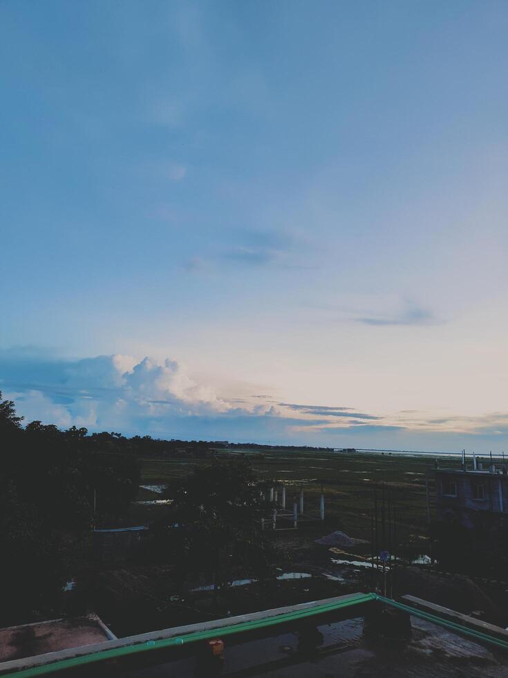 Serene dawn or dusk landscape with human settlement, construction, and marshy fields. Features soft light, fluffy clouds, and a vast sky. photo