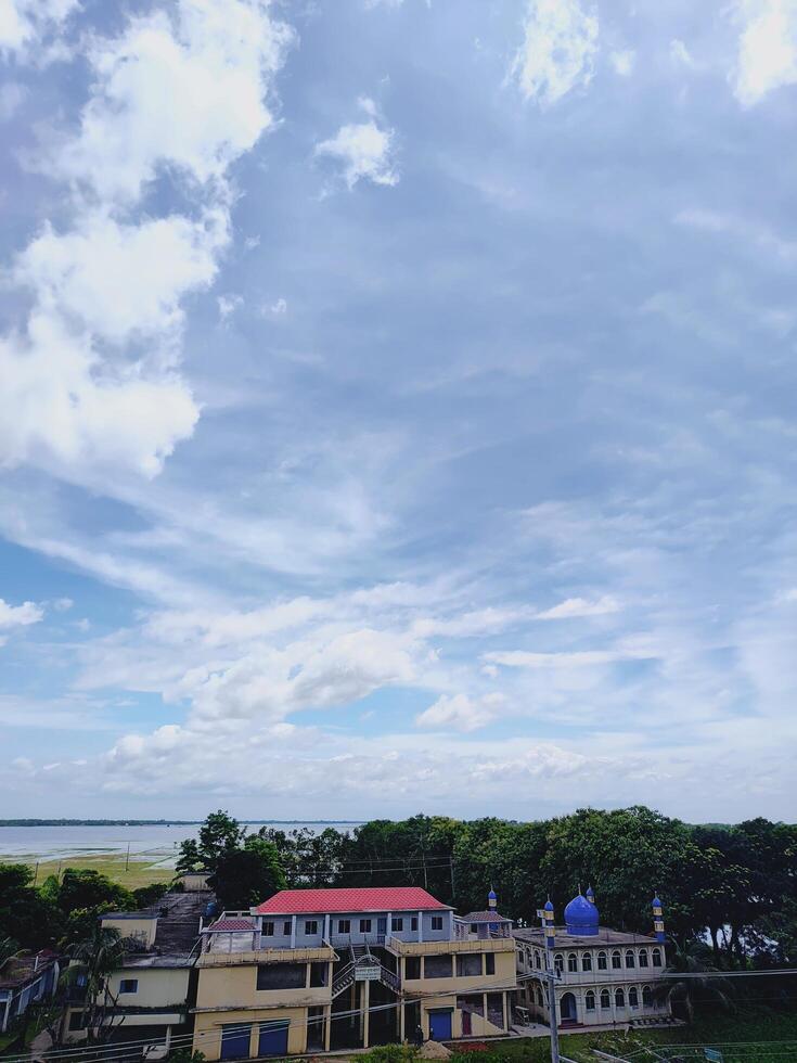 Scenic rural view featuring a mosque and red roofed building surrounded by greenery, with a tranquil river under a cloudy sky. Ideal for nature, culture, and architectural landscapes. photo