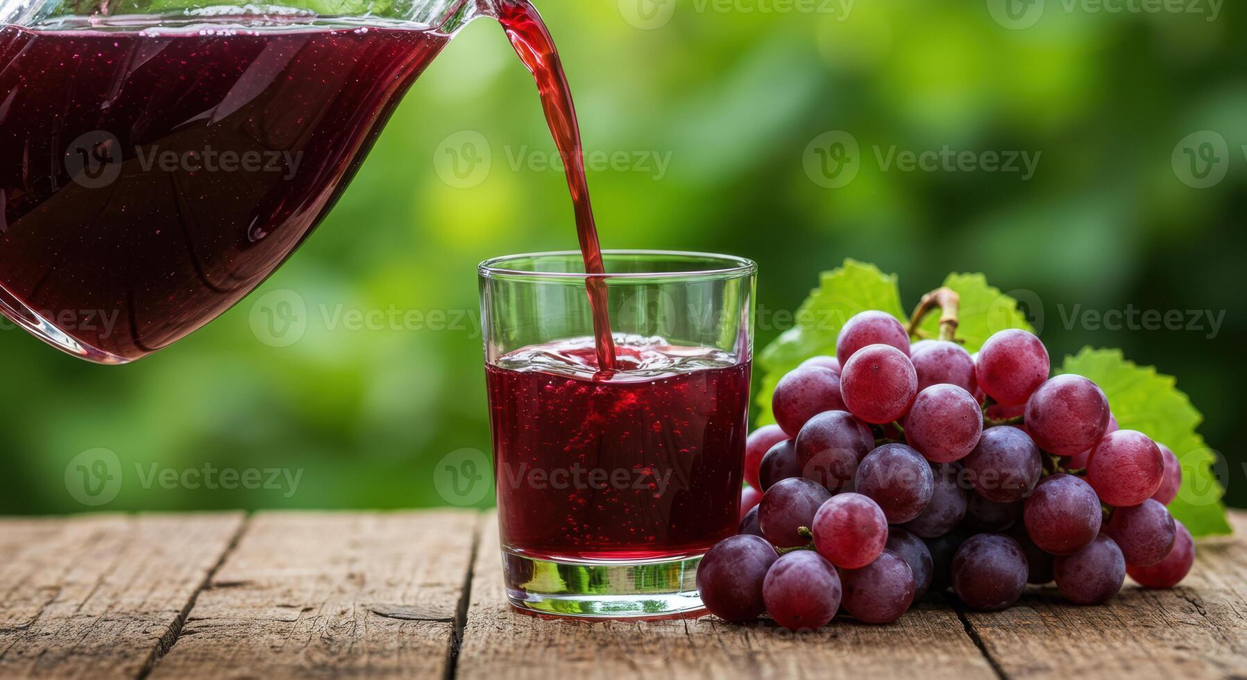 Pouring refreshing grape juice into a clear glass with grapes on a wooden table against green nature backdrop photo