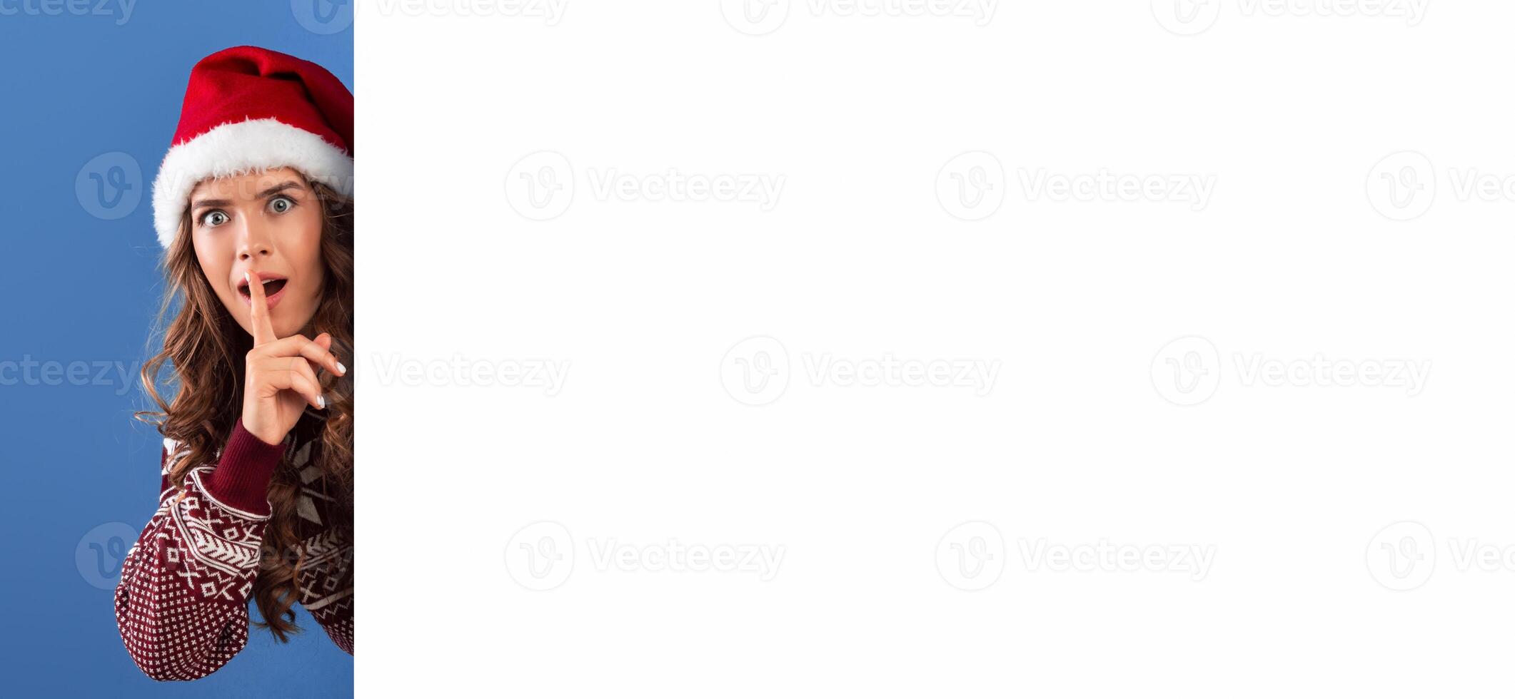 Pretty girl in Christmas outfit peeking out from behind blank poster and making hush gesture, blue background. Space for design. Panorama photo