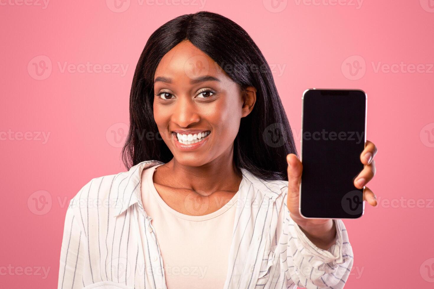 Pretty black woman showing mobile phone with space for your app design, pink studio background. Lovely African American lady holding smartphone with mockup for website. Communications concept photo