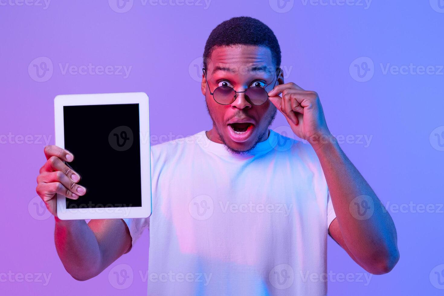 Check This. Amazed African American Guy In Stylish Eyeglasses Holding Digital Tablet With Black Screen, Excited Man Demonstrating Mockup Space For Your App or Website Advertesement Under Neon Light photo