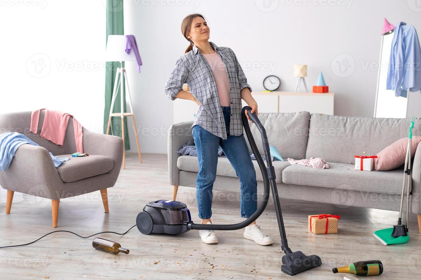 Young woman having back ache from cleaning after party chaos in her apartment, vacuuming floor, empty space. Millennial lady suffering from pain while cleaning dirty room after celebration photo