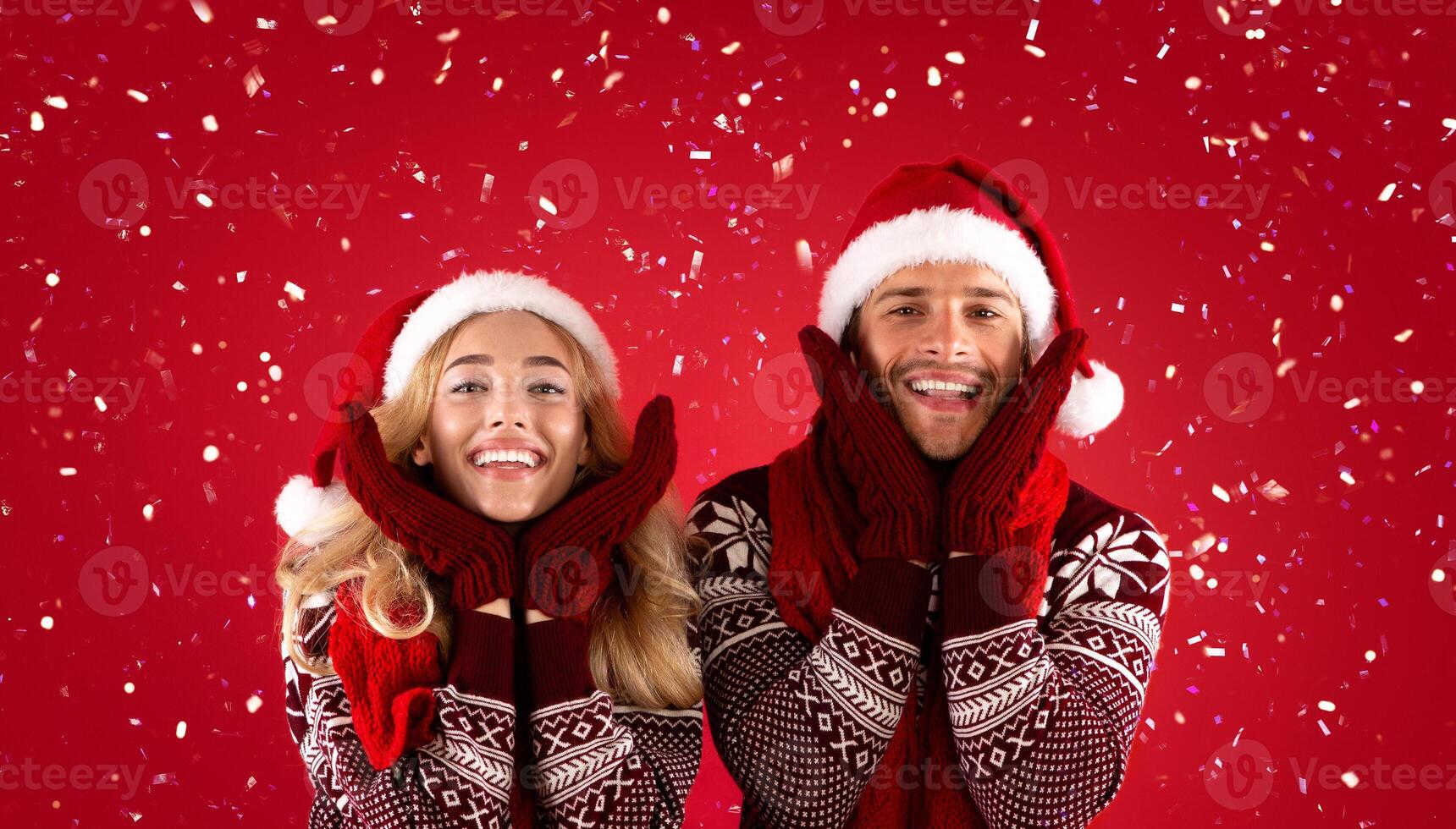 It's Christmas time. Adorable young couple in mittens, Santa hats and scarves on red background photo