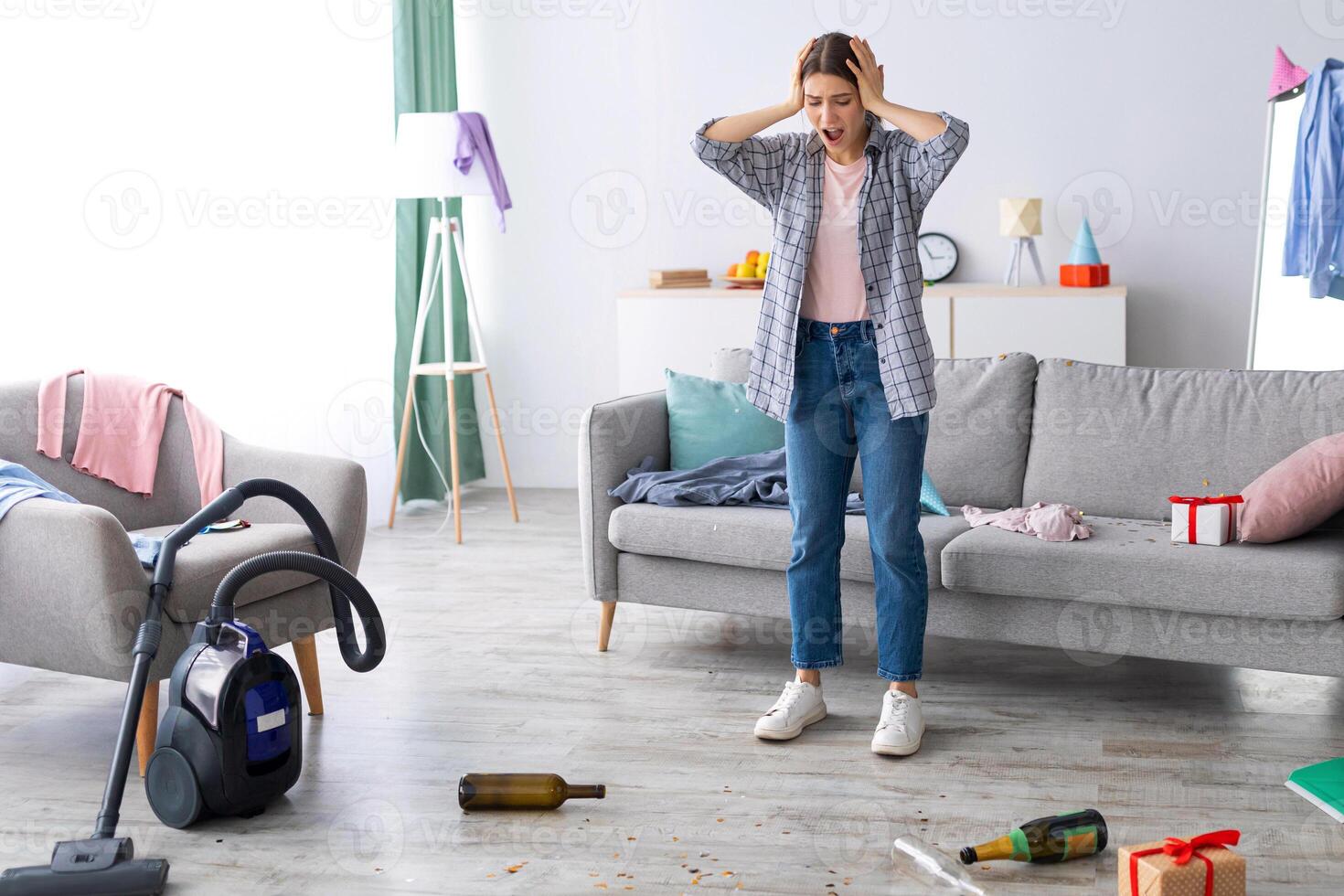 Shocked young woman looking at her messy apartment after party, holding head in terror, not knowing where to start cleaning, copy space. Dismayed female student at dirty college dorm photo