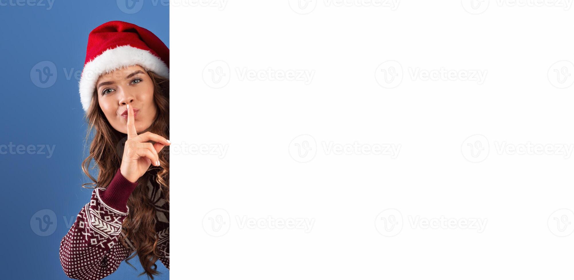 Christmas promotion. Attractive young woman in Santa hat peeking out from behind empty banner and making SILENCE gesture, blue studio background. Space for your design. Panorama photo