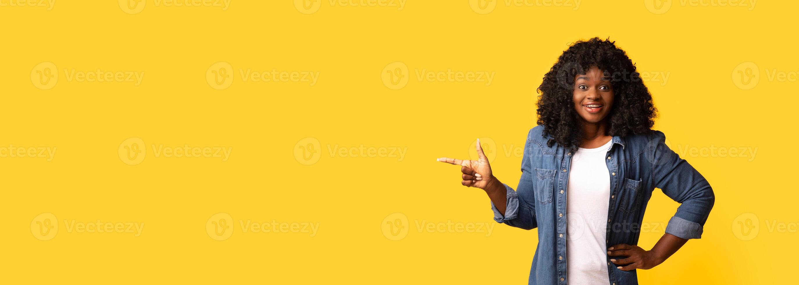 Amazed african american young woman pointing at copy space, yellow studio background. Excited black lady in casual outfut showing empty space for text or advertisement, panorama photo