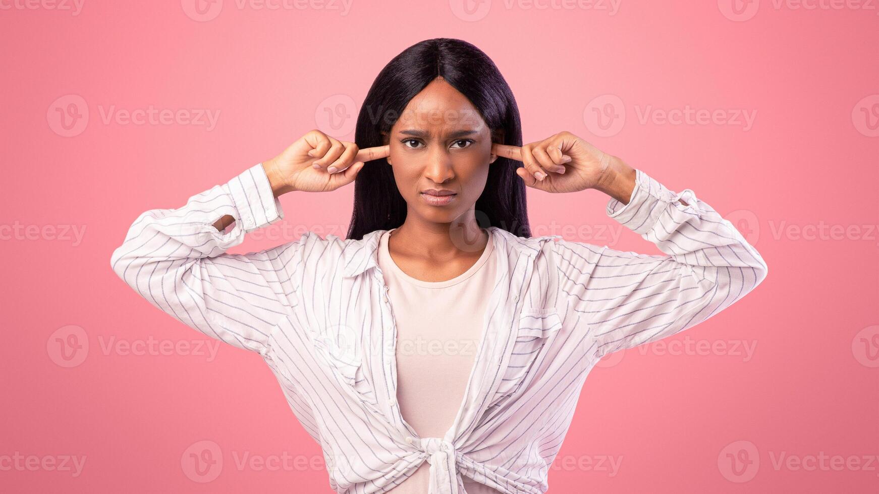Upset black lady closing her ears with fingers, protecting from loud noise on pink studio background. African American woman not willing to listen, ignoring someone, feeling stressed and unhappy photo