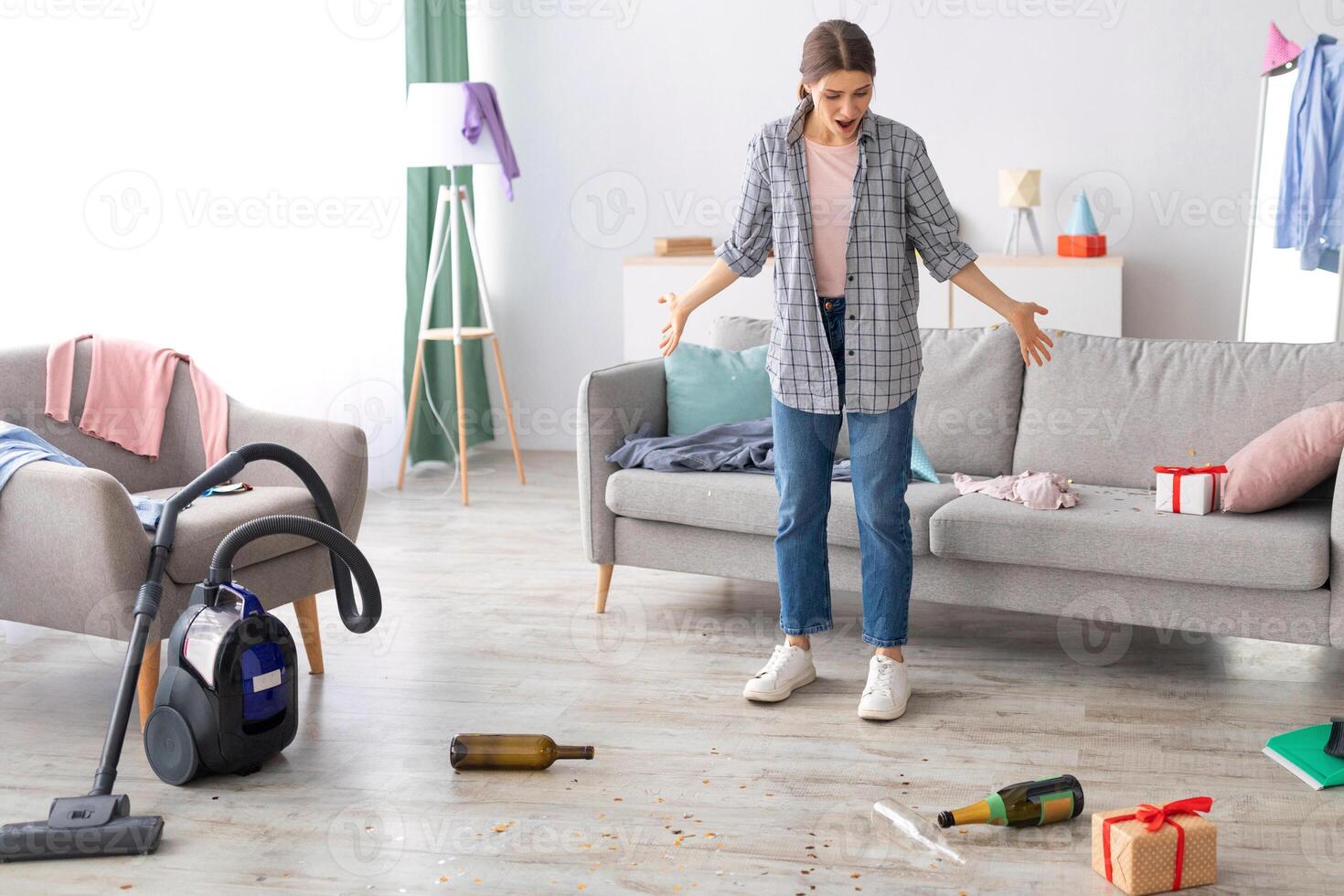 Young woman standing in messy apartment after party, screaming from despair, empty space. Unhappy millennial housewife at dirty room after celebration, rubbish scattered on floor photo