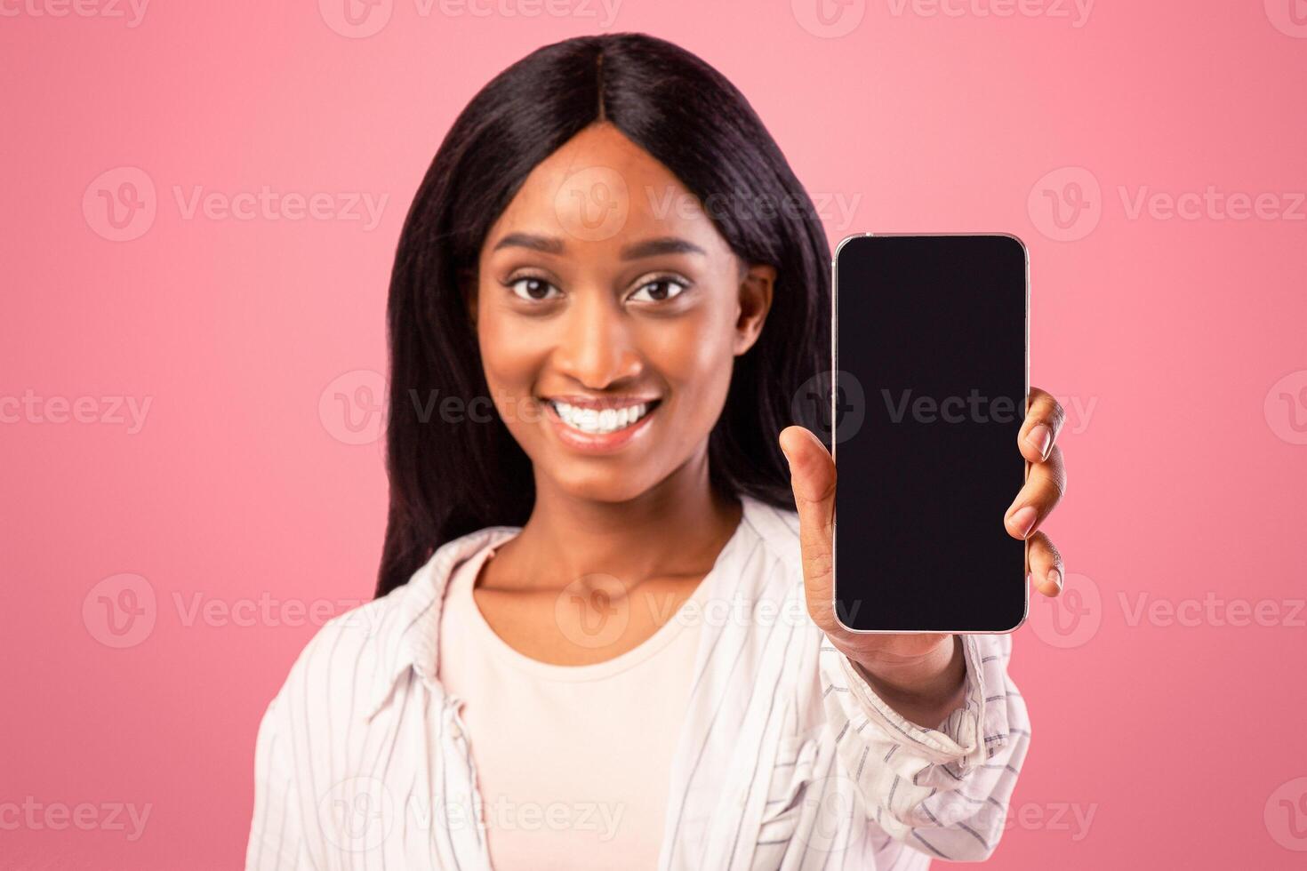 Positive black woman showing cellphone with space for mobile app design on screen over pink studio background, selective focus. Cheerful African American lady holding smartphone with mockup photo