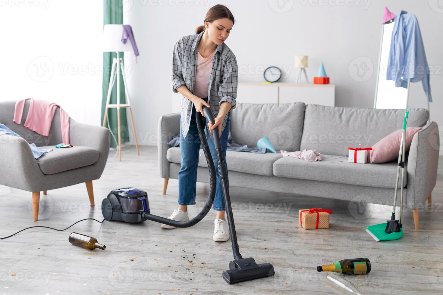 Unhappy young woman vacuuming messy apartment after party, cleaning up chaos after holiday celebration, copy space. Millennial female student doing cleanup at college dorm room photo