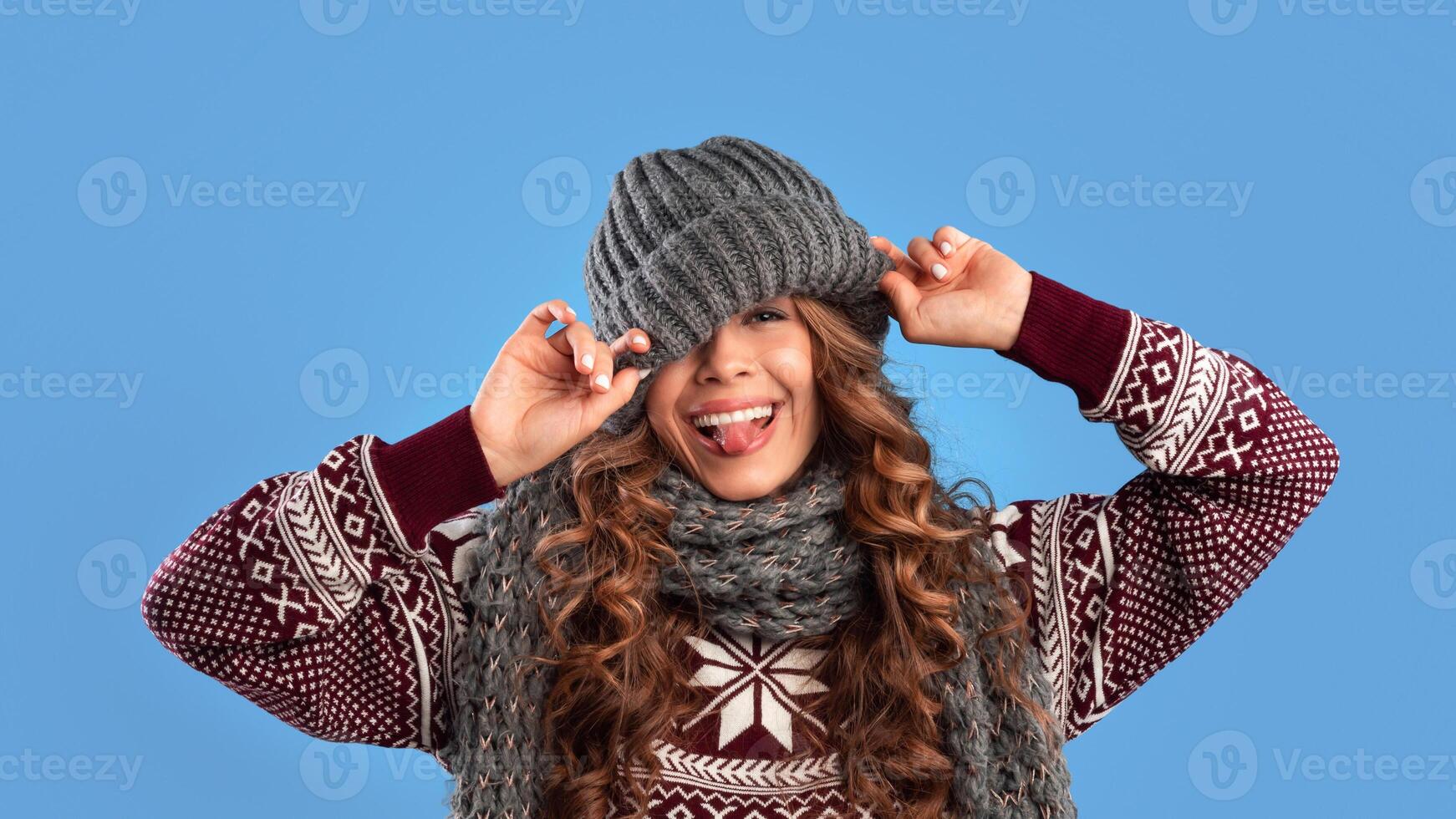Silly young woman in warm winter clothes pulling down knitted beanie hat and sticking out her tongue on blue studio background, panorama. Lovely lady in woolen outfit having fun photo