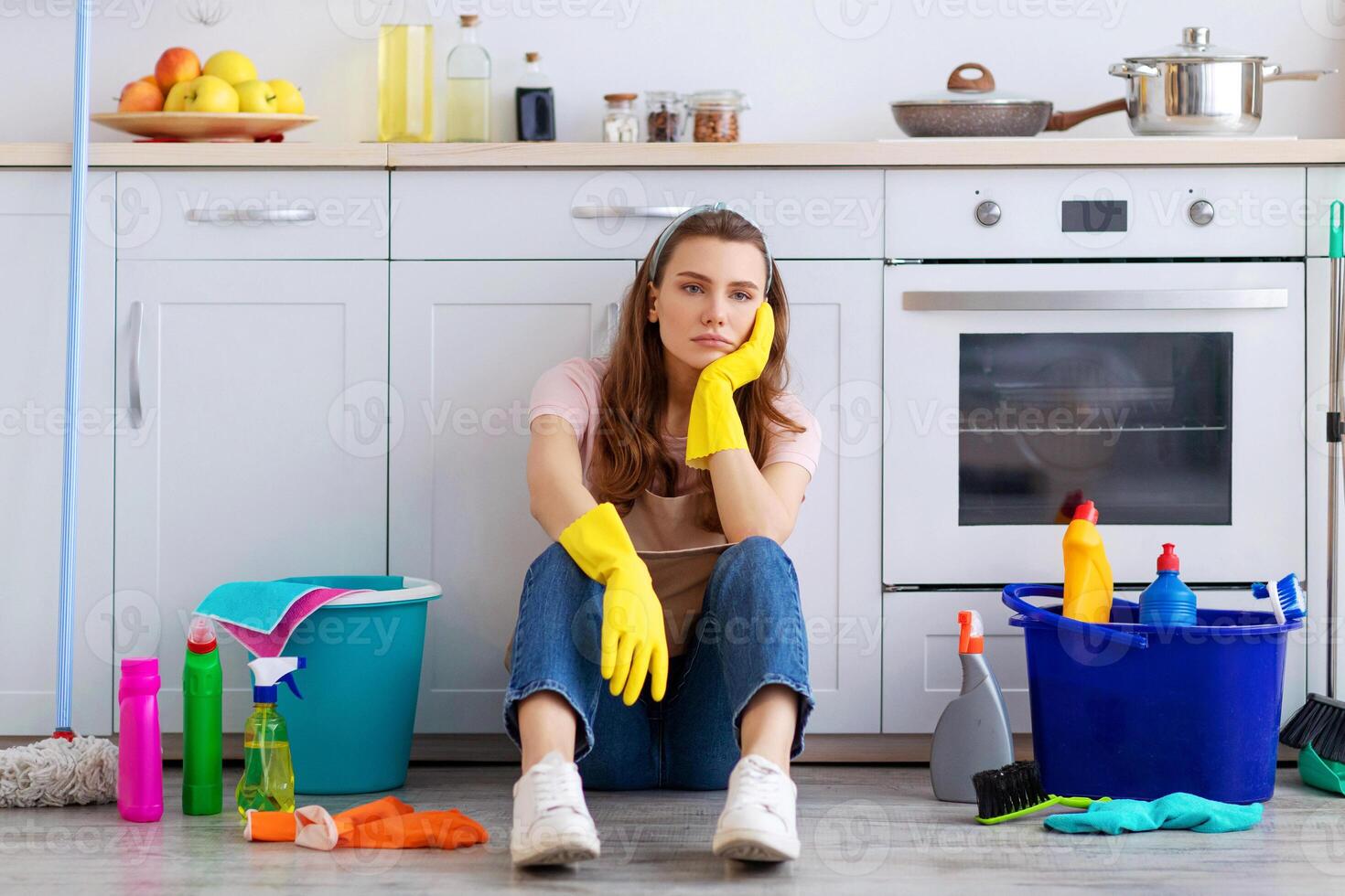 Exhausted young housewife sitting on floor at kitchen, surrounded by cleaning supplies, tired from housework. Overworked woman needing break from domestic duties, cannot stand doing house chores photo