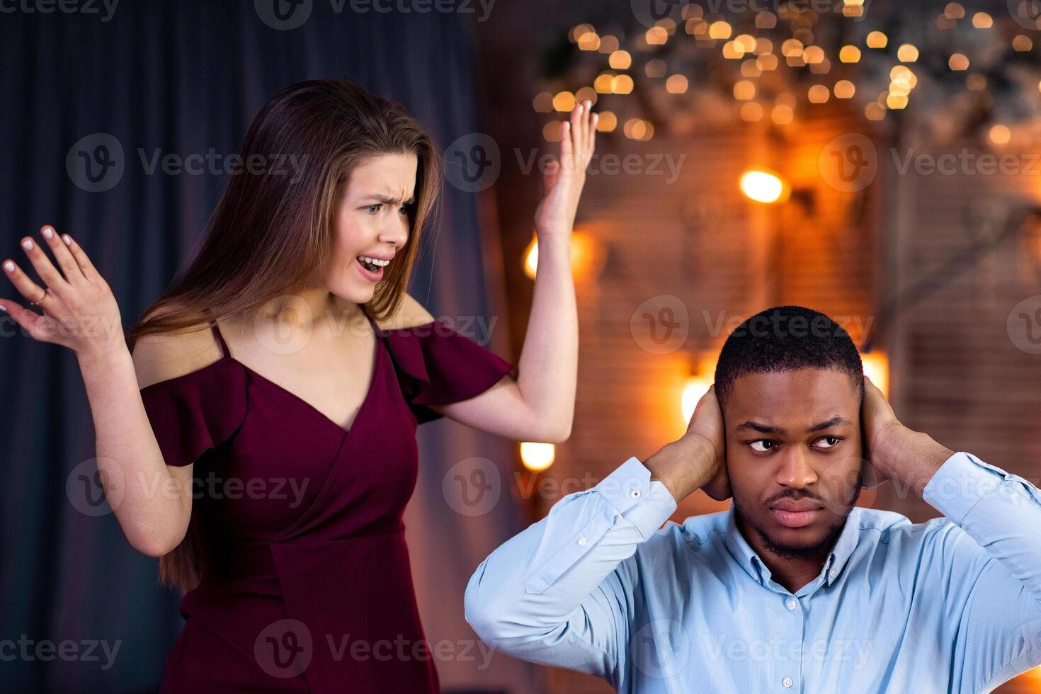 Young Interracial Couple Arguing During Dinner Date In Restaurant. Emotional Woman Shouting At Black Boyfriend, Blaming Him For Cheating And Infidelity, Indifferent Guy Covering Ears With Hands photo