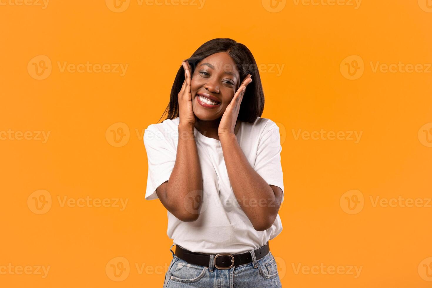 Emotional black lady in white t-shirt smiling and touching her face over orange studio background, happy african american young woman expressing amazement, seeing something exciting, copy space photo