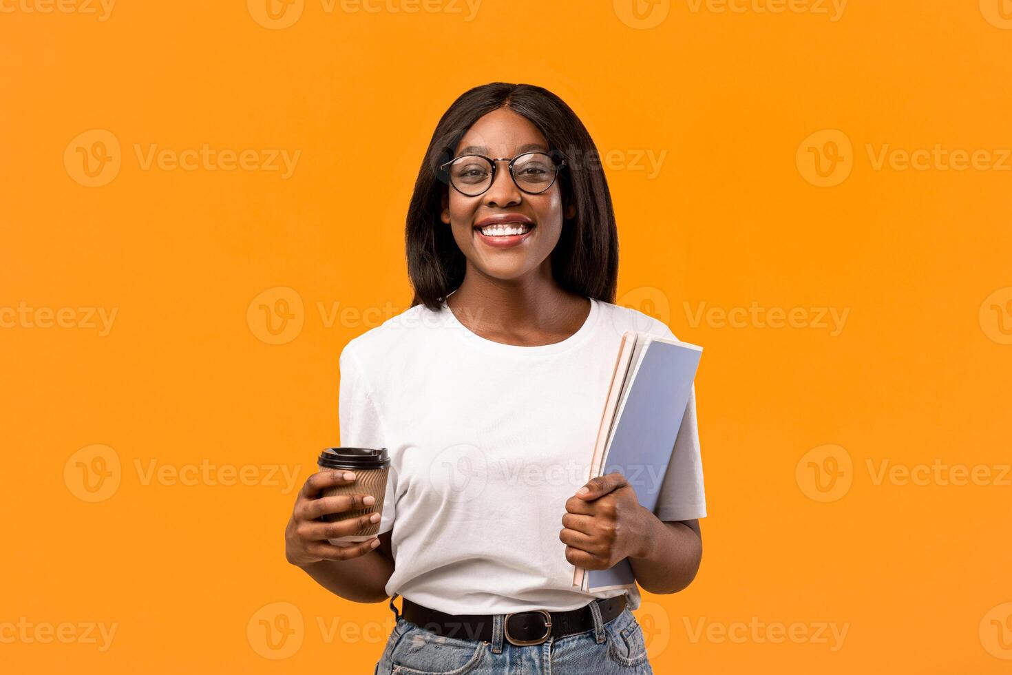 Positive young black lady in glasses student holding notebooks and coffee to go, having break while studying, orange studio background, copy space. Young african american woman on coffee break photo