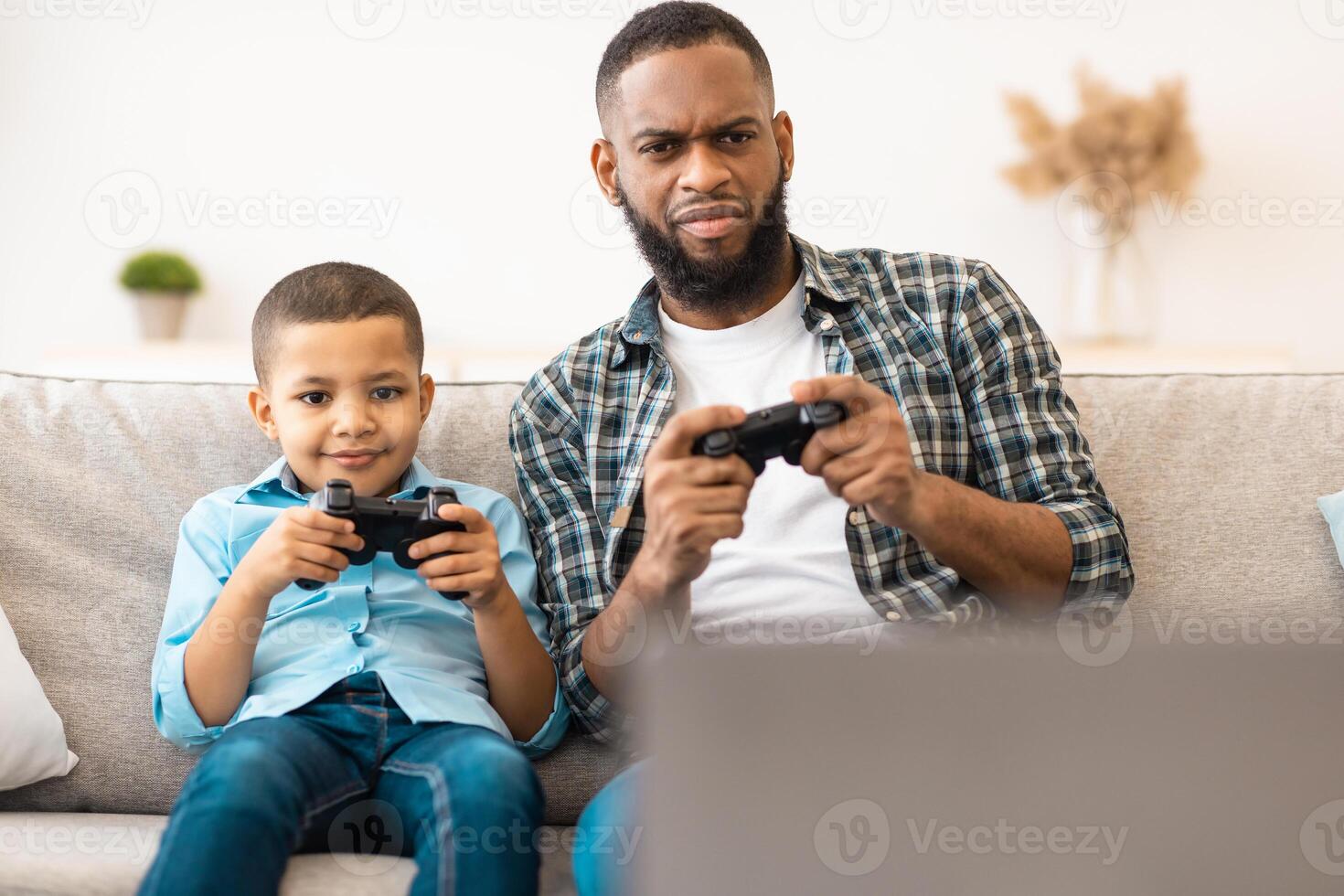 Black Daddy And Son Playing Videogame Having Fun Together Sitting On Sofa At Home. Family Lifestyle, Weekend Leisure, And Computer Gaming Concept. Selective Focus photo