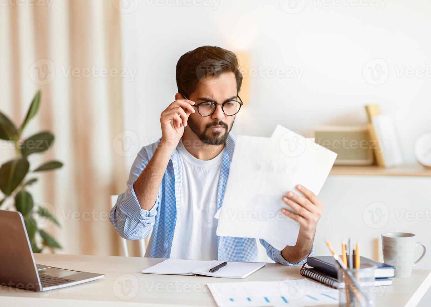 Concerned Arab Businessman In Eyeglasses Reading Financial Documents At Workplace In Office, Eastern Male Entrepreneur Checking Annual Reports While Sitting At Desk With Laptop, Copy Space photo
