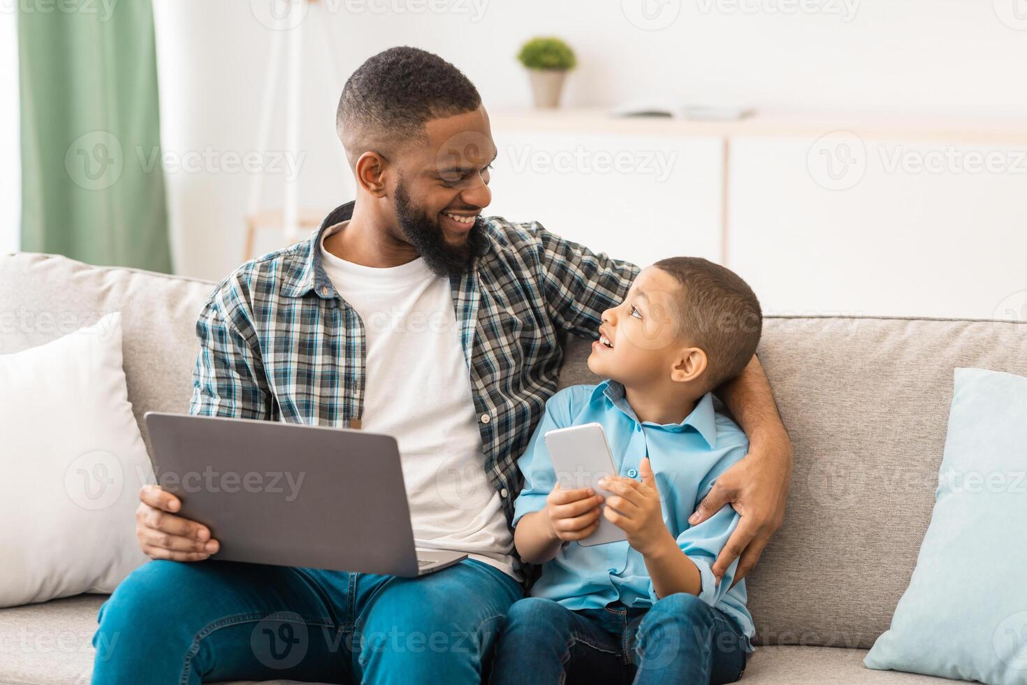 Family Gadgets. Black Daddy And Little Son Using Laptop And Phone Browsing Internet Sitting And Talking Bonding At Home. Weekend With Father, Boy And His Dad Using Computer And Smartphone Together photo