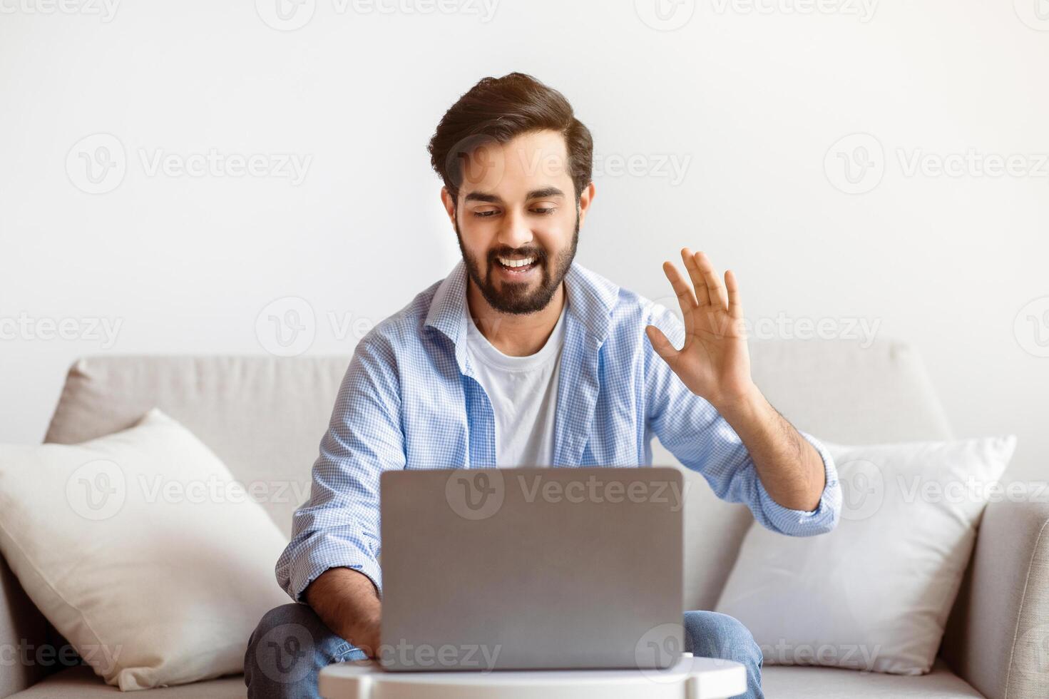 Online Meeting. Cheerful Arab Man Making Call With Laptop At Home, Friendly Eastern Guy Waving Hand At Computer Webcam, Greeting Somebody, Smiling Male Enjoying Distant Communication, Free Space photo