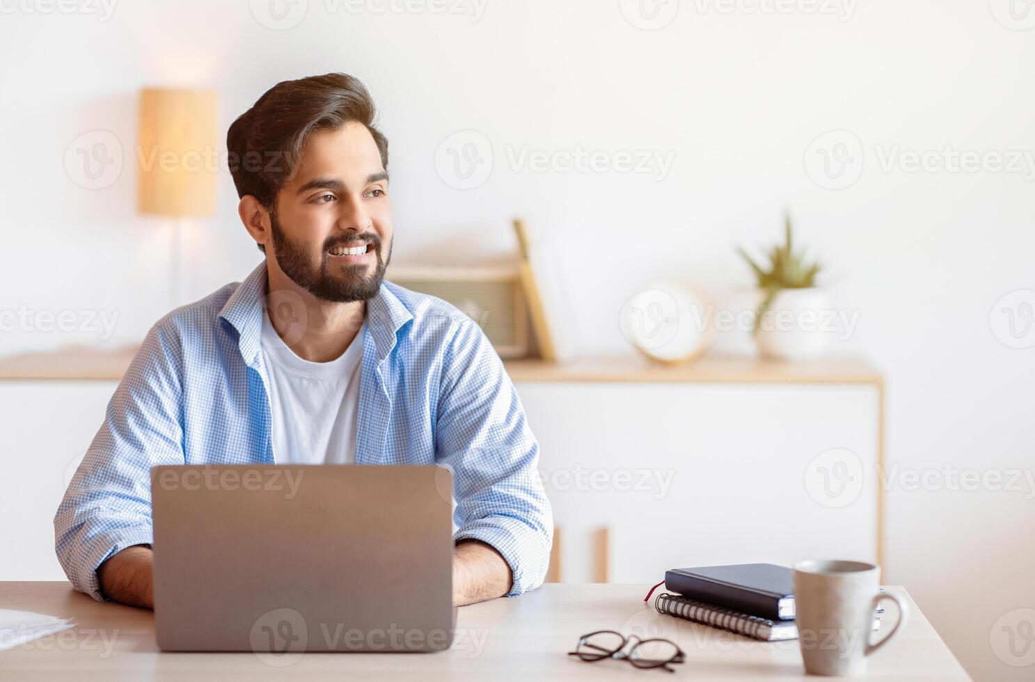 Portrait Of Pensive Arab Man Sitting At Desk With Laptop In Home Office, Looking Away With Glad Face Expression. Smiling Eastern Freelancer Guy Thinking About Successful Business Strategy, Copy Space photo