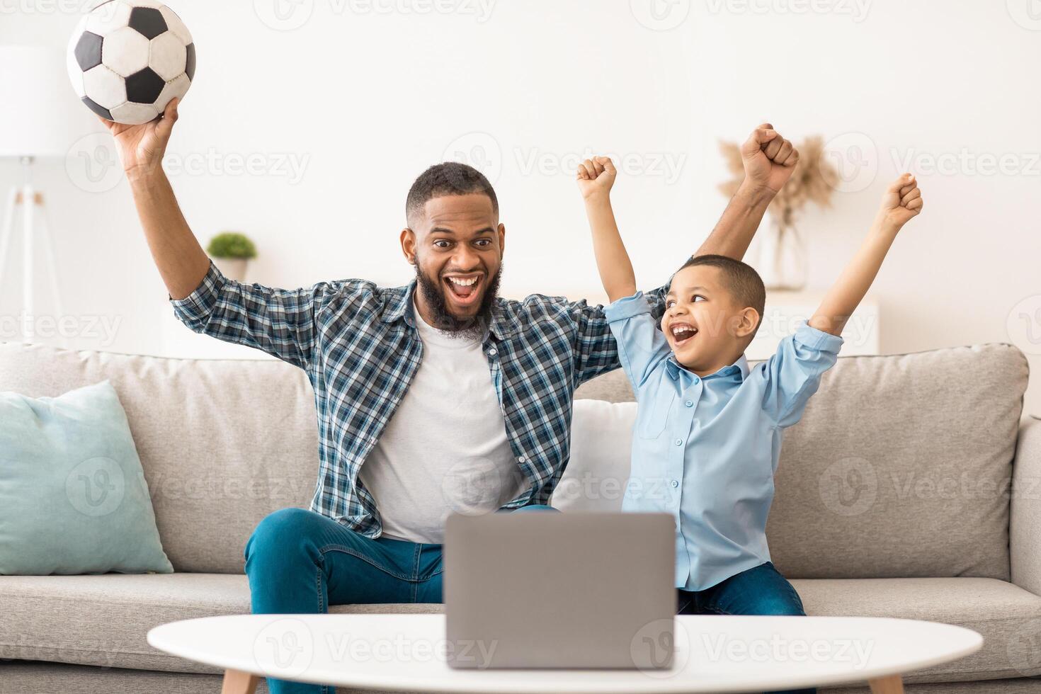 Joyful Black Daddy And Son Watching Sport Game On Laptop Cheering Favorite Soccer Team Sitting On Sofa At Home. Football Fans Watch Online Television And Sports Channels At Computer Concept photo