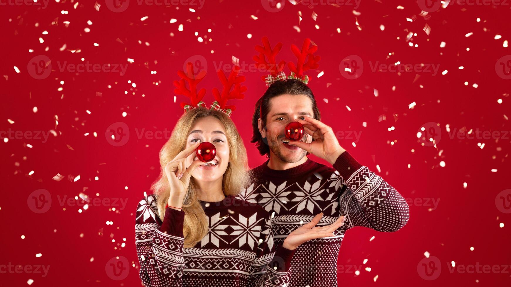 Xmas fun. Silly young couple holding Christmas baubles near their noses on red background photo