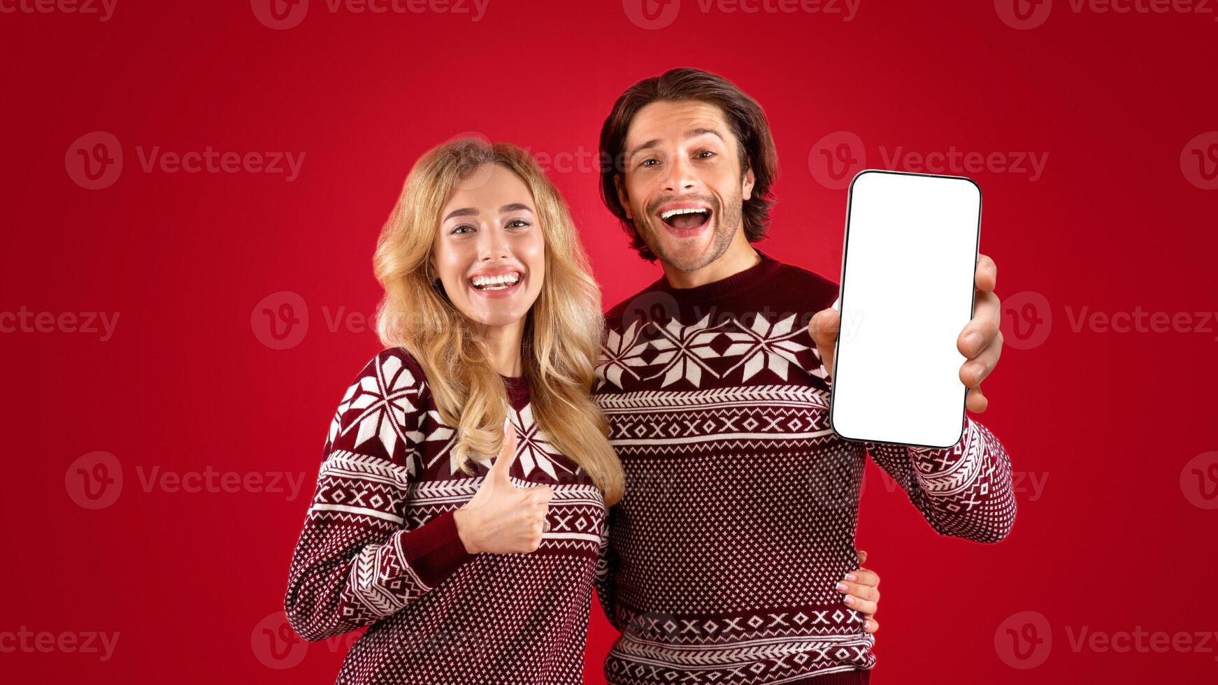 Hapy millennial couple in Christmas sweaters showing thumb up and peace gestures over red background photo