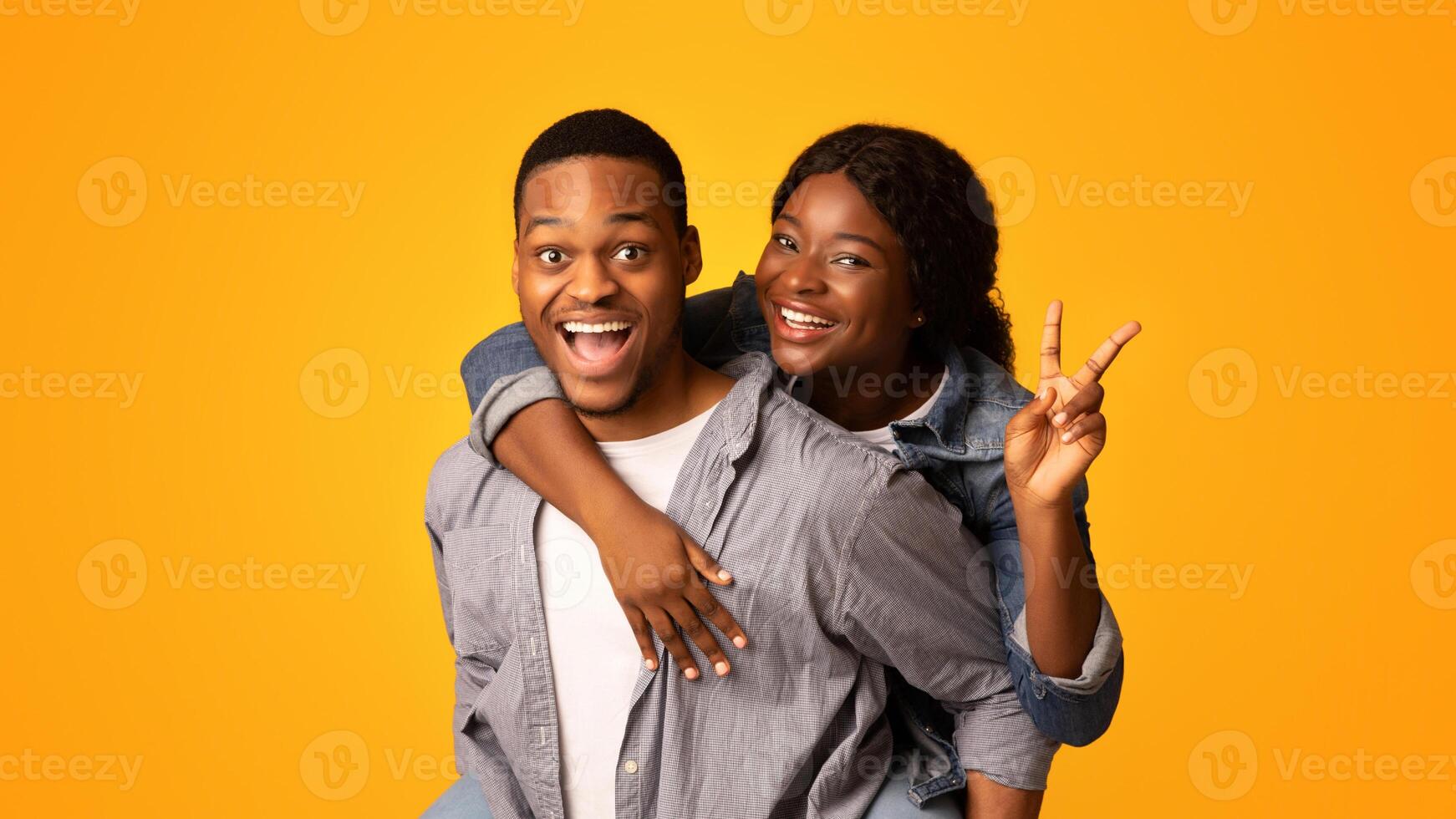 Happy African American Couple Having Fun Together, Black Guy Piggybacking His Cheerful Girlfriend, Joyful Woman Showing Peace Gesture At Camera While Standing Isolated Over Yellow Background photo