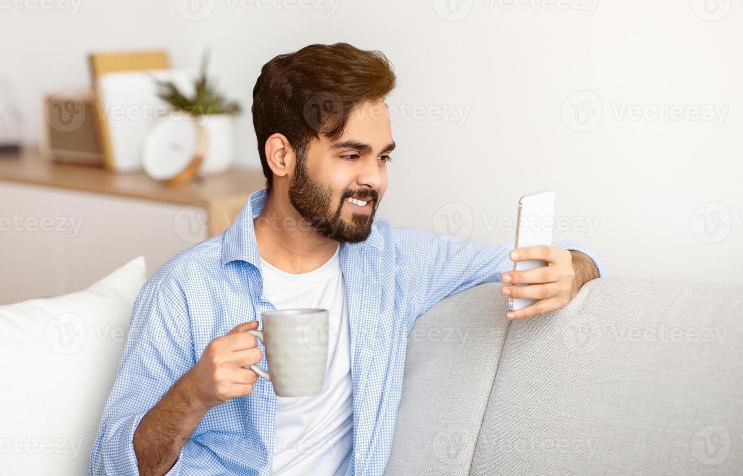 Domestic Leisure. Handsome Eastern Guy Using Smartphone And Drinking Coffee At Home While Resting On Couch In Living Room, Watching Videos Or Making Call, Enjoying Weekend Pastime, Free Space photo