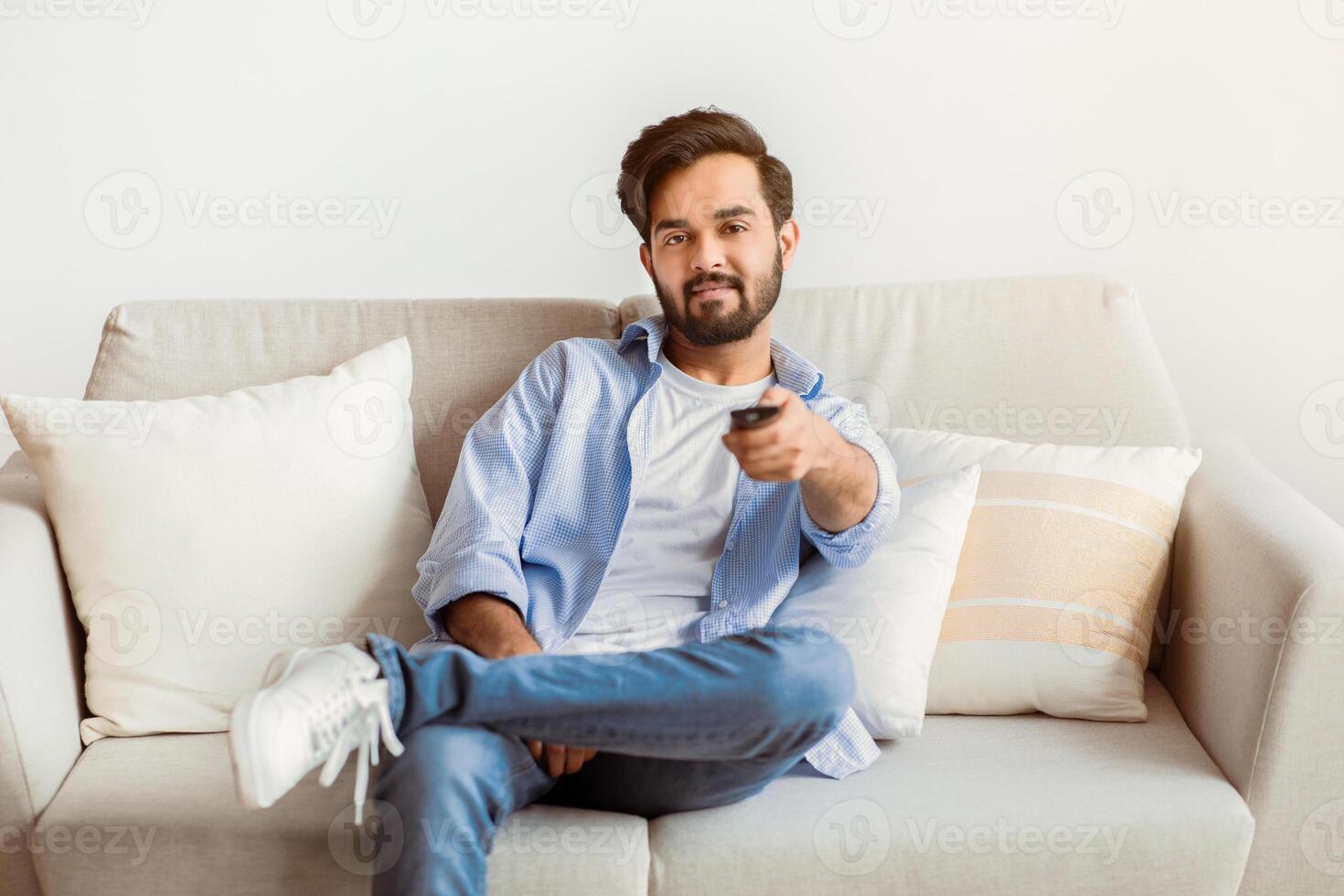 Smiling Young Middle Eastern Guy Watching TV At Home. Millennial Arab Man Relaxing On Couch In Living Room And Switching Channels With Remote Controller, Enjoying Domestic Weekend, Copy Space photo