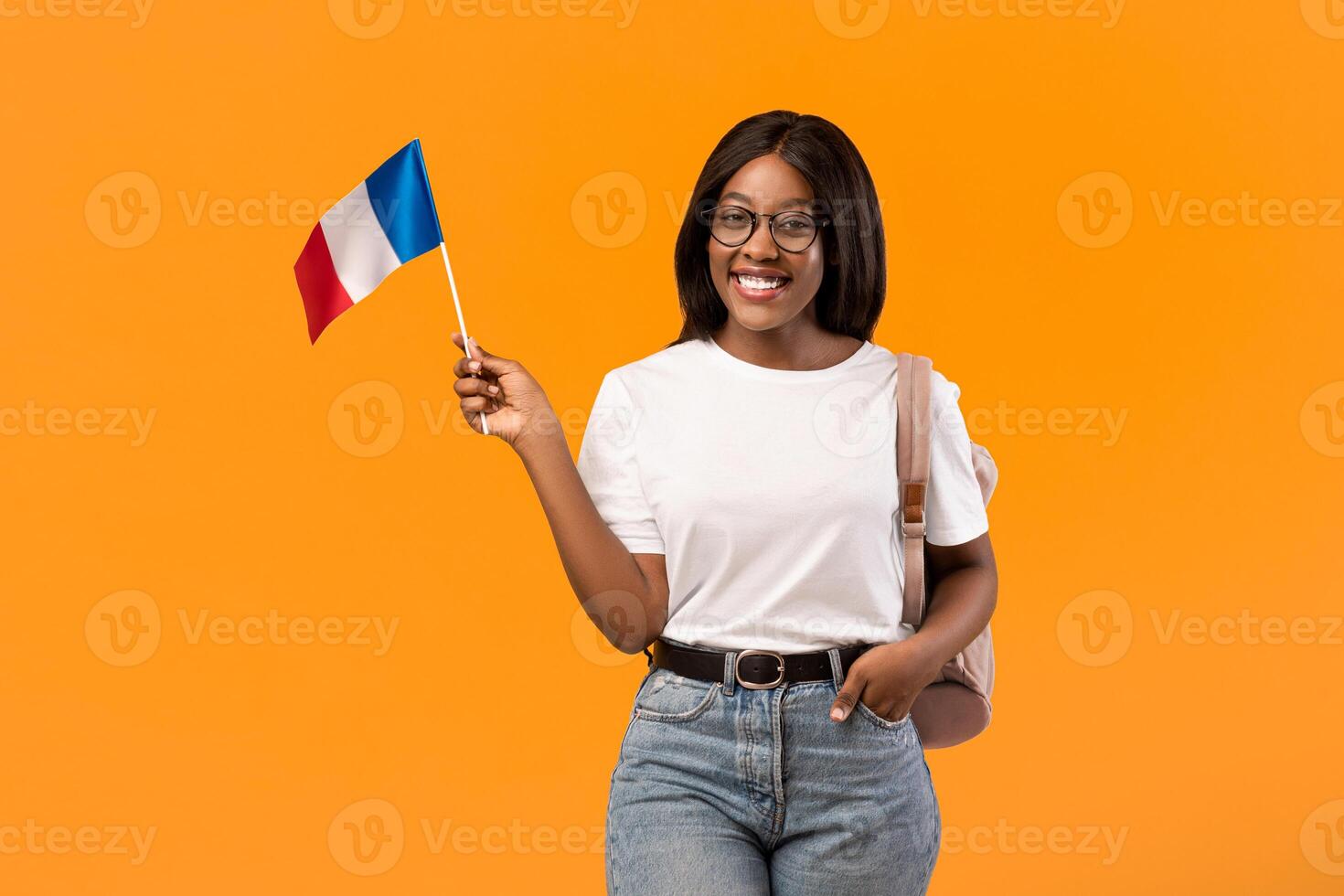 Happy young black woman student in white t-shirt showing french flag, orange studio background, copy space. Smiling african american lady with backpack and glasses showing flag of France photo