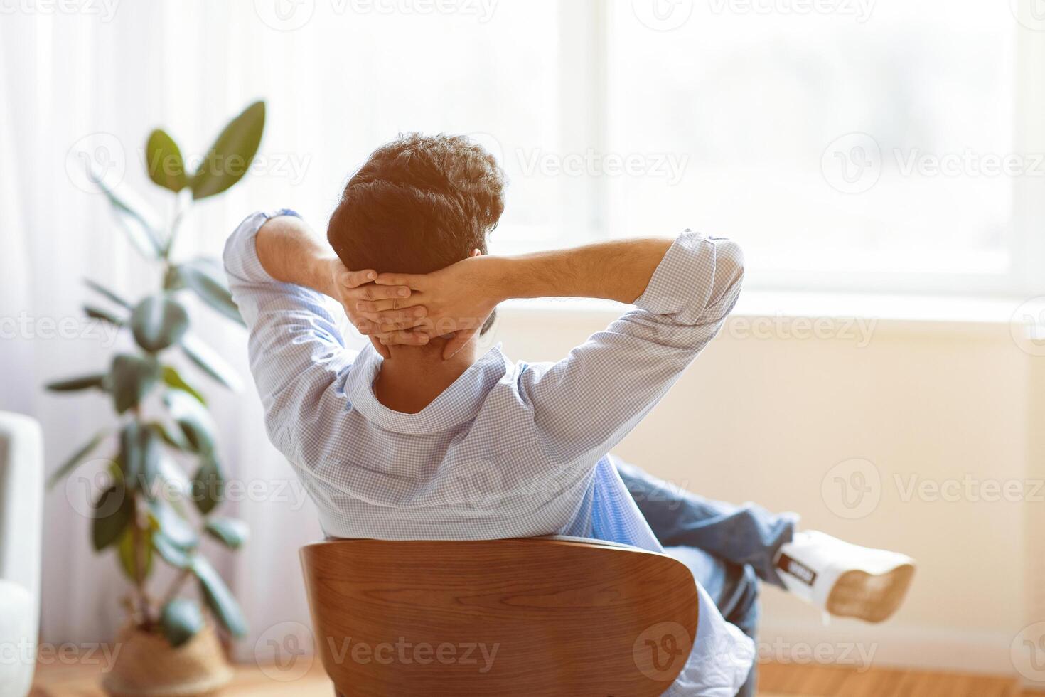 Rear view of unrecognizable man relaxing on chair in cozy interior at home with hands behind head, dark haired male leaning back and looking to window, enjoying lazy domestic day off, copy space photo