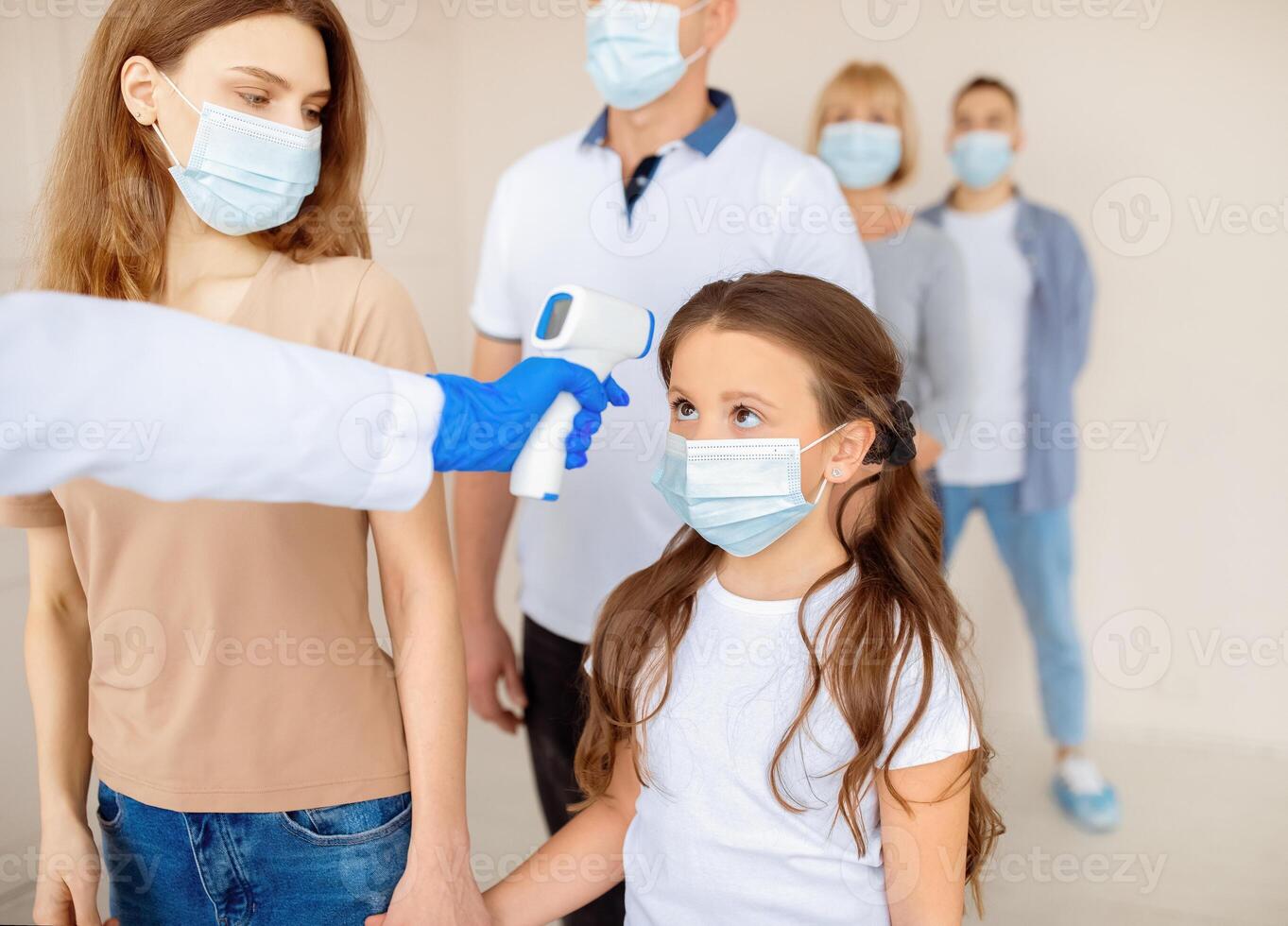 Nurse checking little girl's and her mom's temperature with infrared thermometer in queue for coronavirus vaccine injection at clinic. People waiting for covid-19 immunization. Healthcare concept photo
