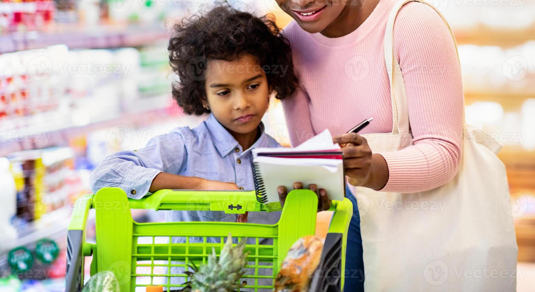 Millennial black mother and her adorable daughter checking grocery list in notebook while shopping at supermarket. Happy African American family purchasing food together at store photo
