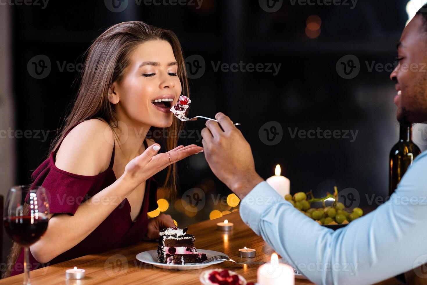 Try it. Cheerful mixed couple feeding each other with dessert, having fun at romantic date in restaurant, elegant multiracial lovers enjoying celebrating valentine's day or anniversary together photo