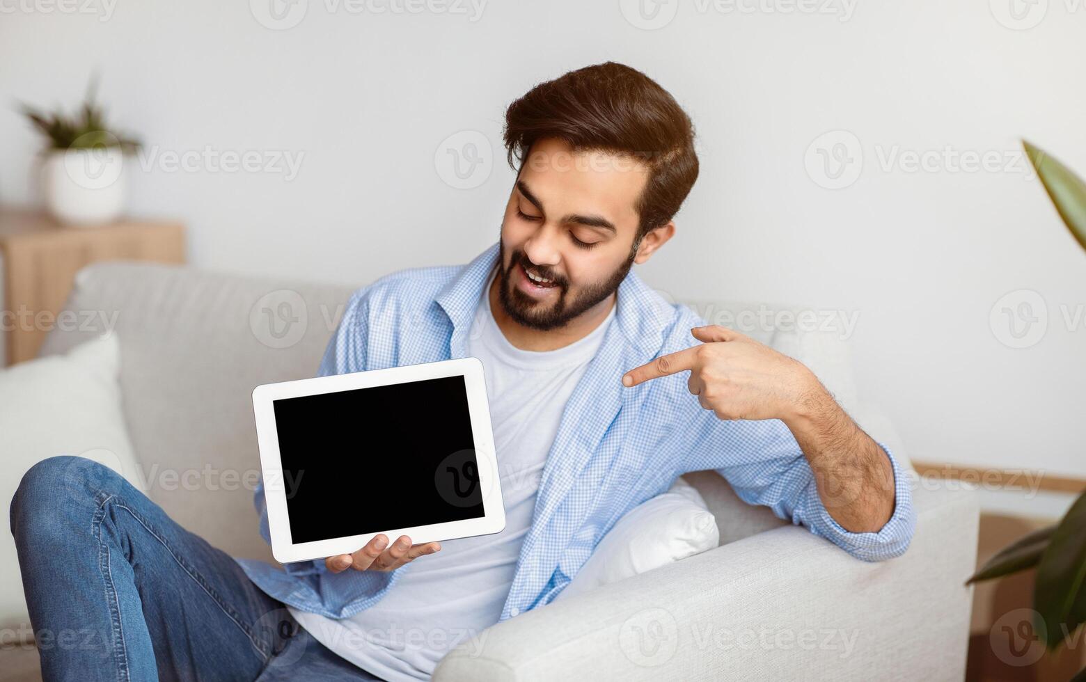 Cheerful Young Eastern Man Pointing At Digital Tablet With Black Screen At Home. Millennial Arab Man Demonstrating Copy Space For Your App Or Website Design, Relaxing On Couch In Living Room, Mockup photo