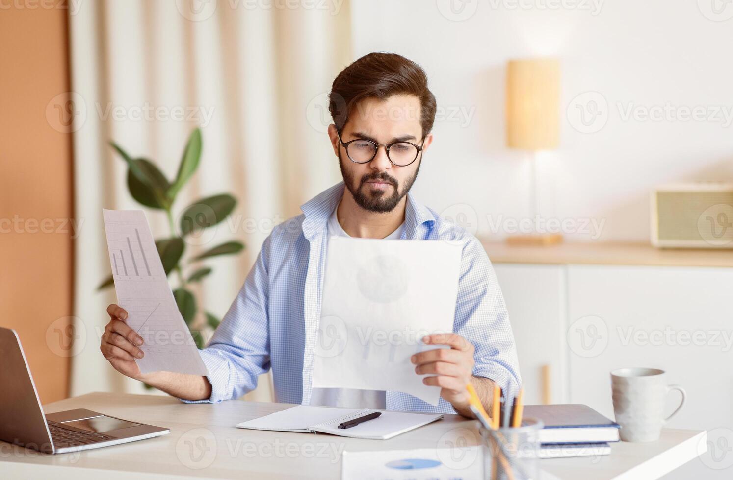 Handsome Eastern Male Entrepreneur Reading Financial Papers While Sitting At Desk In Office, Focuses Arab Businessman In Eyeglasses Checking Annual Reports At Workplace, Free Space photo