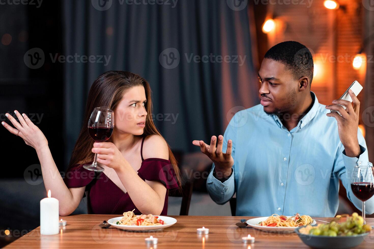 Young Woman Complaining About Her Black Boyfriend Talking On Cellphone During Romantic Date In Restaurant, Feeling Annoyed And Bored. Interracial Couple Having Problems In Relationship, Free Space photo