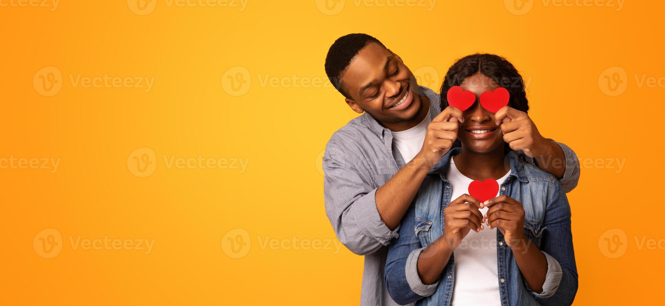 Romance Concept. , Romantic African American Guy Covering Eyes Of His Girlfriend With Red Paper Hearts, Happy Black Couple Having Fun On Valentine's Day, Isolated On Yellow Background, Free Space photo