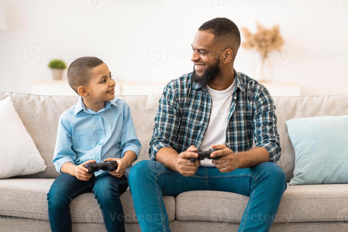 Happy Black Daddy And His Kid Son Playing Games Smiling Each Other Sitting With Game Controllers At Home. Family Having Fun On Weekend, Play Videogame Bonding Together. photo