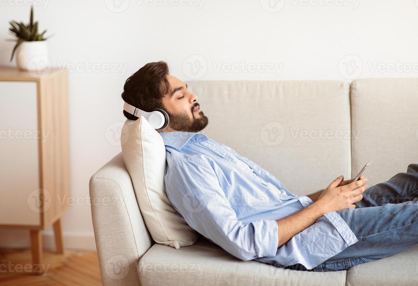 Young Arab Man In Wireless Headphones Relaxing On Couch With Smartphone, Calm Eastern Man Listening Favorite Music Or Audiobook While Resting On Sofa In Living Room, Side View With Free Space photo