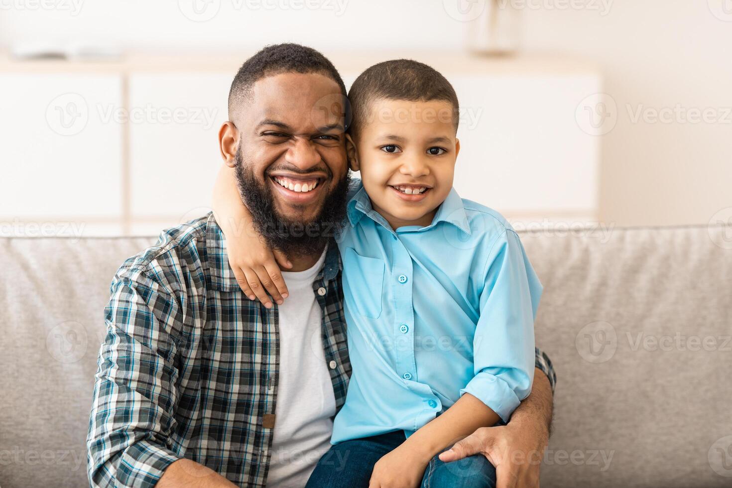 Father's Day. Happy African American Daddy And His Little Son Hugging Posing Together Embracing And Smiling To Camera Sitting On Couch At Home. Portrait Of Happy Dad And Kid Boy. Fatherhood Concept photo